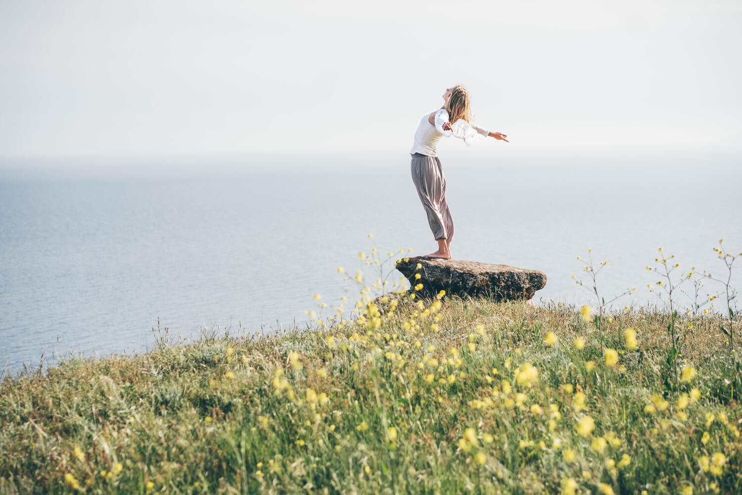 A person stands on a rock with arms outstretched, overlooking the sea. They are surrounded by green grass and yellow wildflowers on a cliff edge under a clear sky.